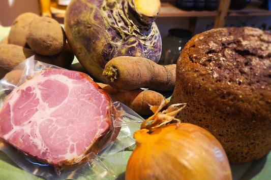 Rustikale Kochbox aus Kraftpapier gefüllt mit einer gelben Steckrübe, Kartoffeln, Möhren, einem Stück Kassler, einem Glas Senf und frischem Schwarzbrot auf ländlichem Hintergrund